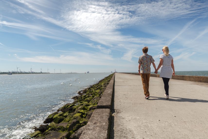 Jong stel wandelt over pier van Hoek van Holland. Wind turbines zichtbaar in achtergrond.