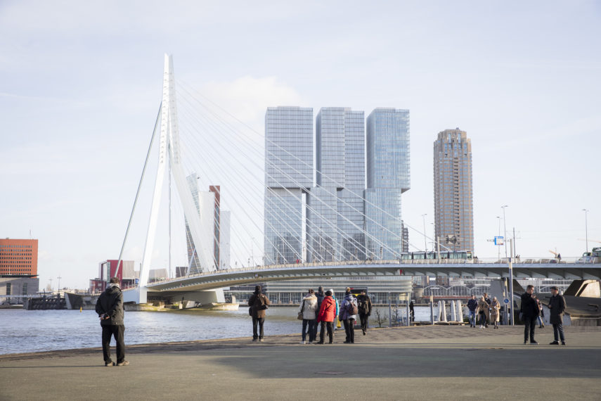 Mensen op de kade voor de Erasmusbrug en op de achtergrond De Rotterdam. 