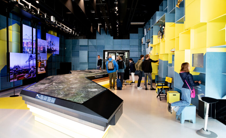 We see people visiting the Port Pavilion, learning about the port of Rotterdam. A port host is showing things to a visitor.