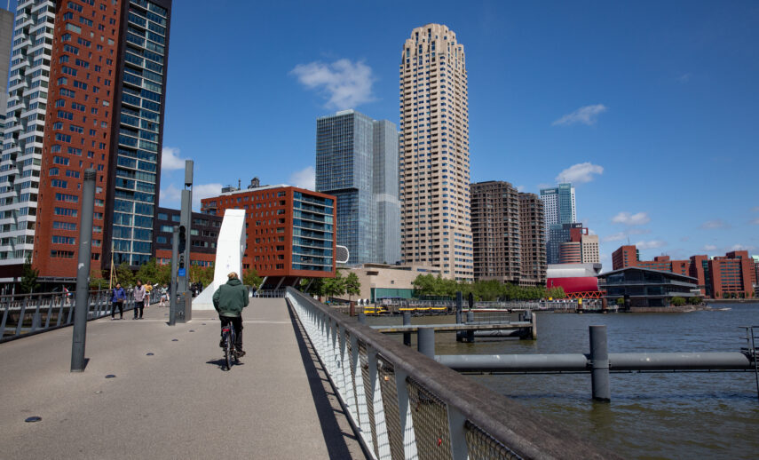 We see a cyclist and some pedestrians crossing the Rijnhavenbridge. In this shot we can also see these buildings: Montevideo, De Rotterdam, Floating Office, New Orleans and in the background Luxor Theatre.