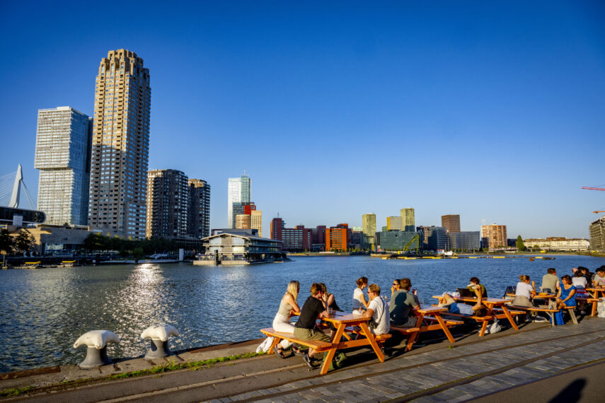 Een foto van mensen die genieten van een zomeravond op de Kop van Zuid. Enkele van de iconische Rotterdamse gebouwen zoals De Rotterdam, New Orleans en Floating Office bevinden zich op de achtergrond