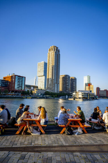 Een foto van mensen die genieten van een zomeravond op de Kop van Zuid. Op de achtergrond zijn Rotterdamse architectuuriconen te zien, waaronder de gebouwen Erasmusbrug, New Orleans, De Rotterdam, AKD en Deloitte.