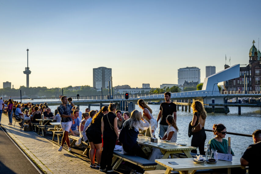 Mensen genieten van een zomeravond op de Kop van Zuid, bij de Fenix ​​Food Factory. Op de achtergrond zijn enkele Rotterdamse iconen te zien, zoals de Euromast, Hotel New York en het Erasmus MC ziekenhuis