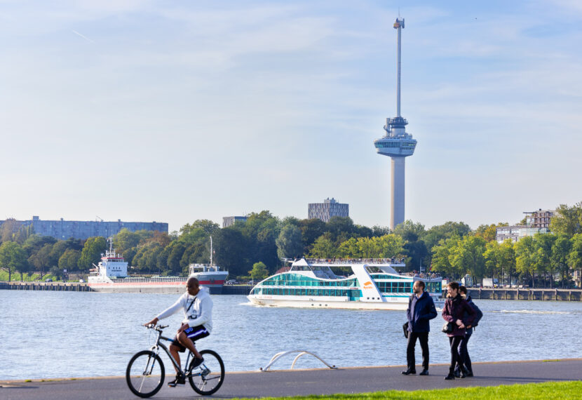 Een foto van mensen die langs de Maas lopen en waar de Spido-boot op vaart. Op de achtergrond is de iconische Euromast van Rotterdam te zien.