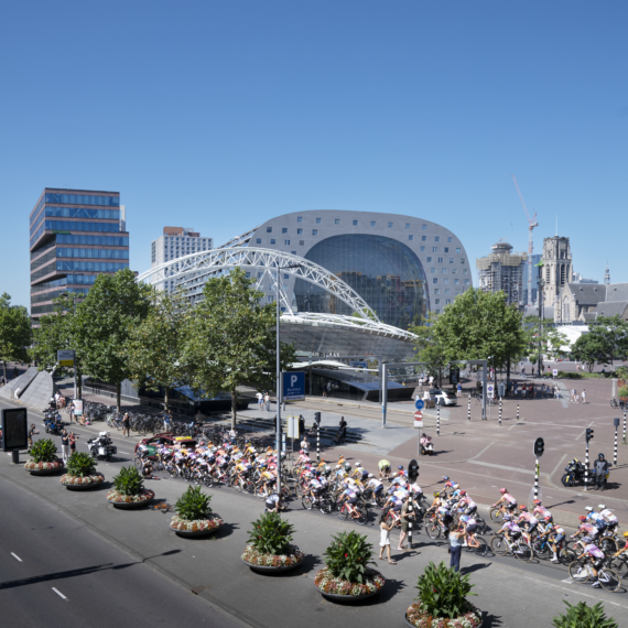 De wielrensters van Tour de France Femmes rijden langs de Markthal. Toeschouwers staan ook de kant van de weg de fietser aan te moedigen. Op de achtergrond is de Laurenskerk te zien onder een heldere, blauwe lucht.
Foto is gemaakt door 