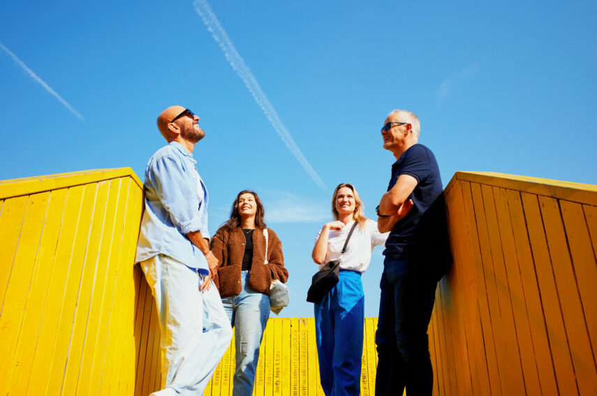 Een groep mensen staat gezellig te kletsen op de gele loopbrug Luchtsingel onder een heldere blauwe lucht. 