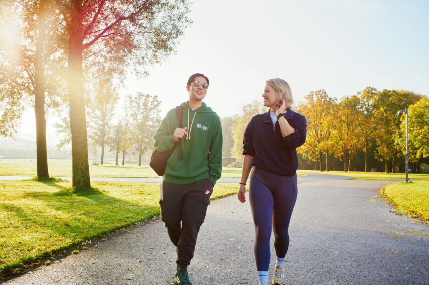 Twee jonge mensen in sportieve kleding wandelen ontspannen over een geasfalteerd pad in het groene Zuiderpark. Ze lachen en zijn in gesprek met elkaar. De zon schijnt fel tussen de bomen door, wat een warme en energieke sfeer geeft.