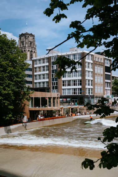 Midden in het centrum van Rotterdam ligt RiF010, waar een surfer meegaat op de golf. Langs de kant staan toeschouwers te kijken, met op de achtergrond stadsgebouwen en de Laurenskerk. 