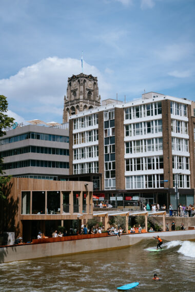 Midden in het centrum van Rotterdam ligt RiF010, waar de ene surfer net een golf heeft gepakt en de ander er nog op meesurft. Langs de kant staan toeschouwers te kijken, met op de achtergrond stadsgebouwen en de Laurenskerk. 

