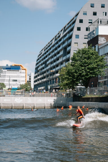 Een surfer gaat mee op de golf bij Rif010., terwijl op de achtergrond de iconische Markthal en de Bibliotheek op de Binnenrotte zichtbaar zijn. 