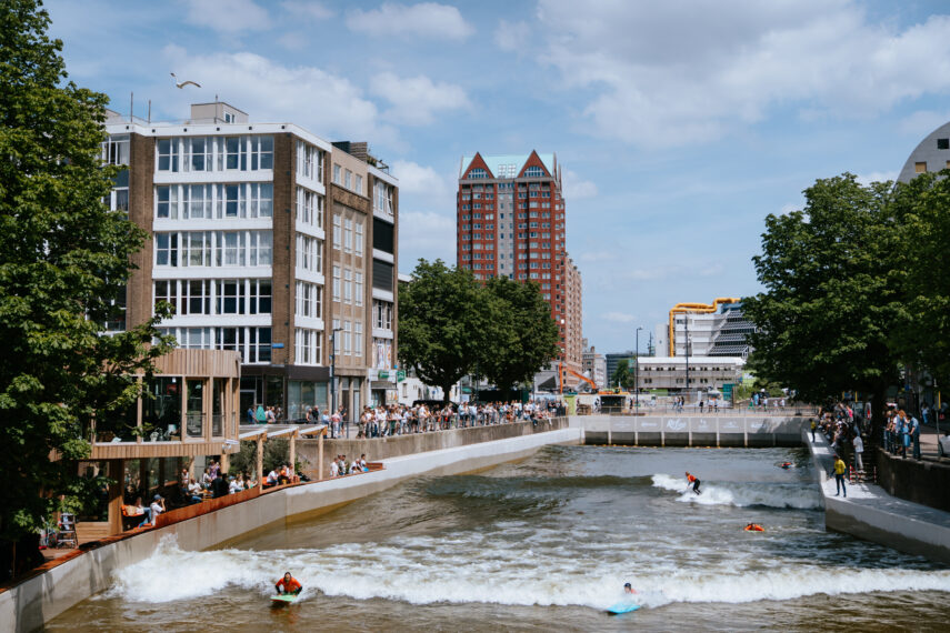 Op een blauwe, lichtbewolkte dag in het hart van Rotterdam trotseren surfers bij RiF010 de golven, terwijl toeschouwers langs de waterkant genieten van het spektakel. Op de achtergrond zijn stadsgebouwen, de iconische bibliotheek aan de Binnenrotte en de Markthal te zien. 