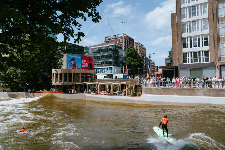 In het water bij RiF010 liggen twee surfers, waarvan één net een golf meepakt. Langs de kant staan toeschouwers aandachtig mee te kijken. Op de achtergrond zijn stadsgebouwen het zichtbaar, en aan de linkerkant de Surfbar & Kitchen en de surfshop van RiF010.