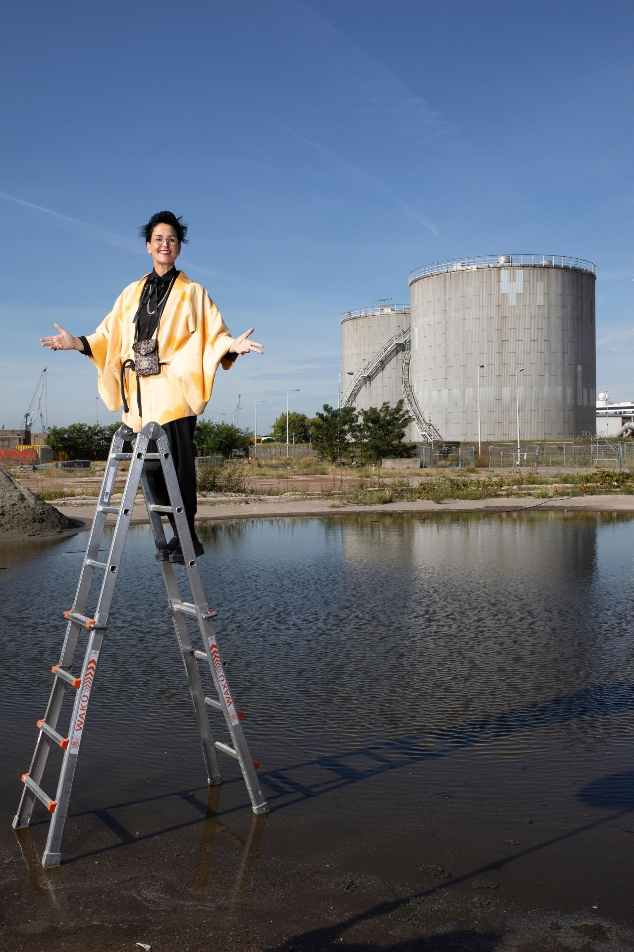 Persoon in gele kimono staat op ladder boven water, met industriële tanks op de achtergrond.