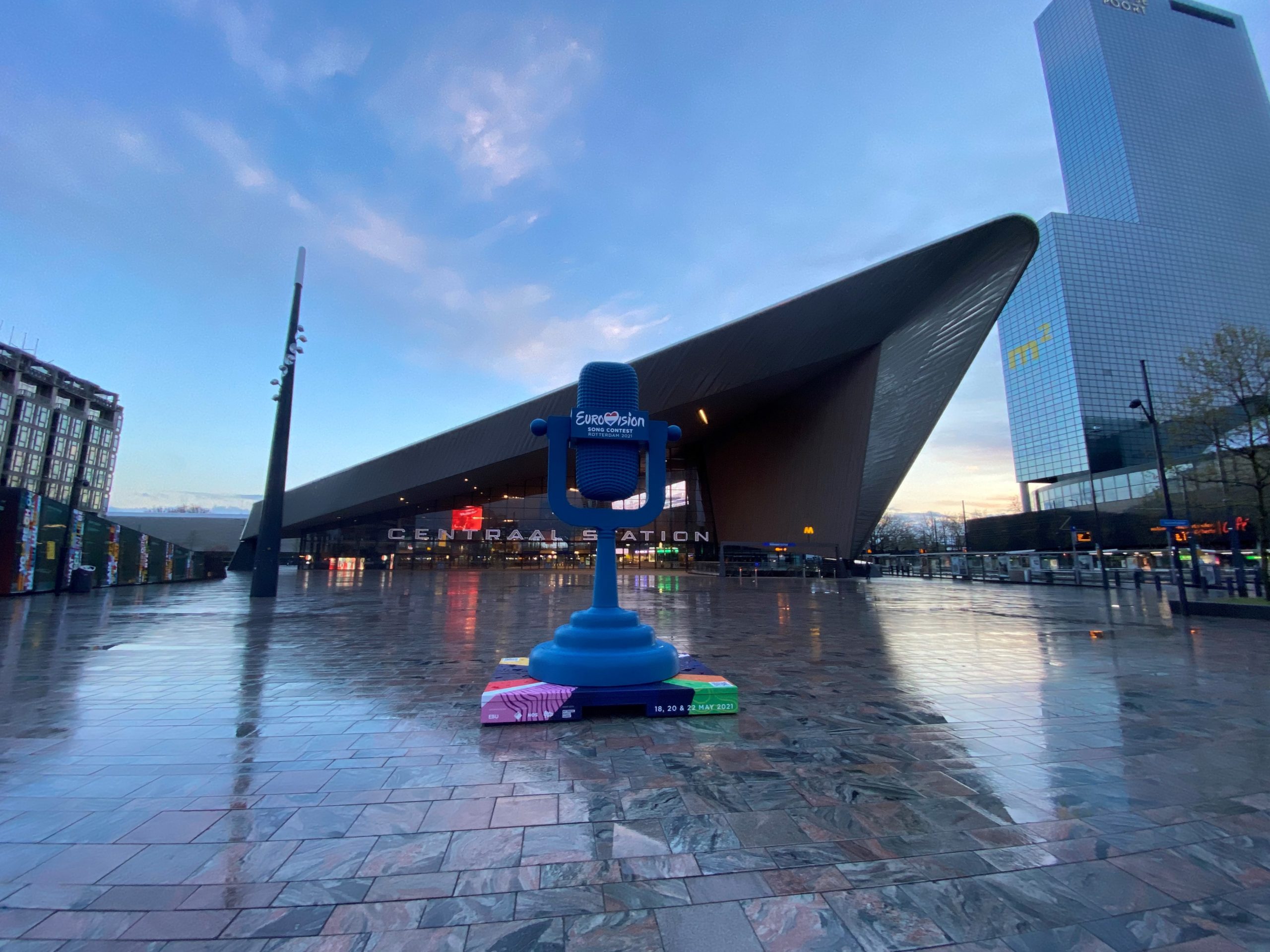 Blauwe Eurovisie-trofee voor Rotterdam Centraal Station bij schemering, met moderne gebouwen op de achtergrond.