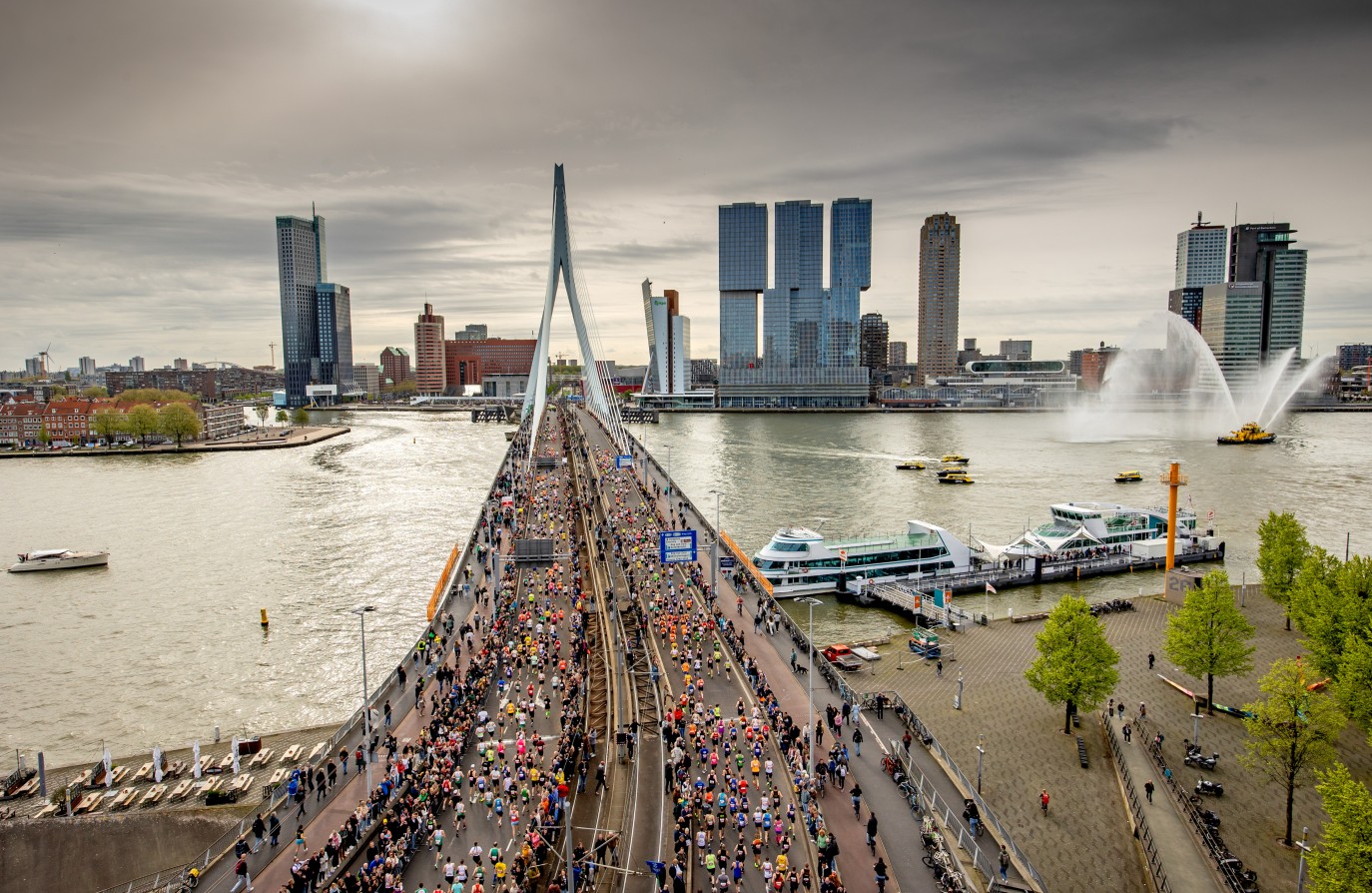 Mensen rennen over de Erasmusbrug in Rotterdam tijdens een marathon, met skyline op achtergrond.