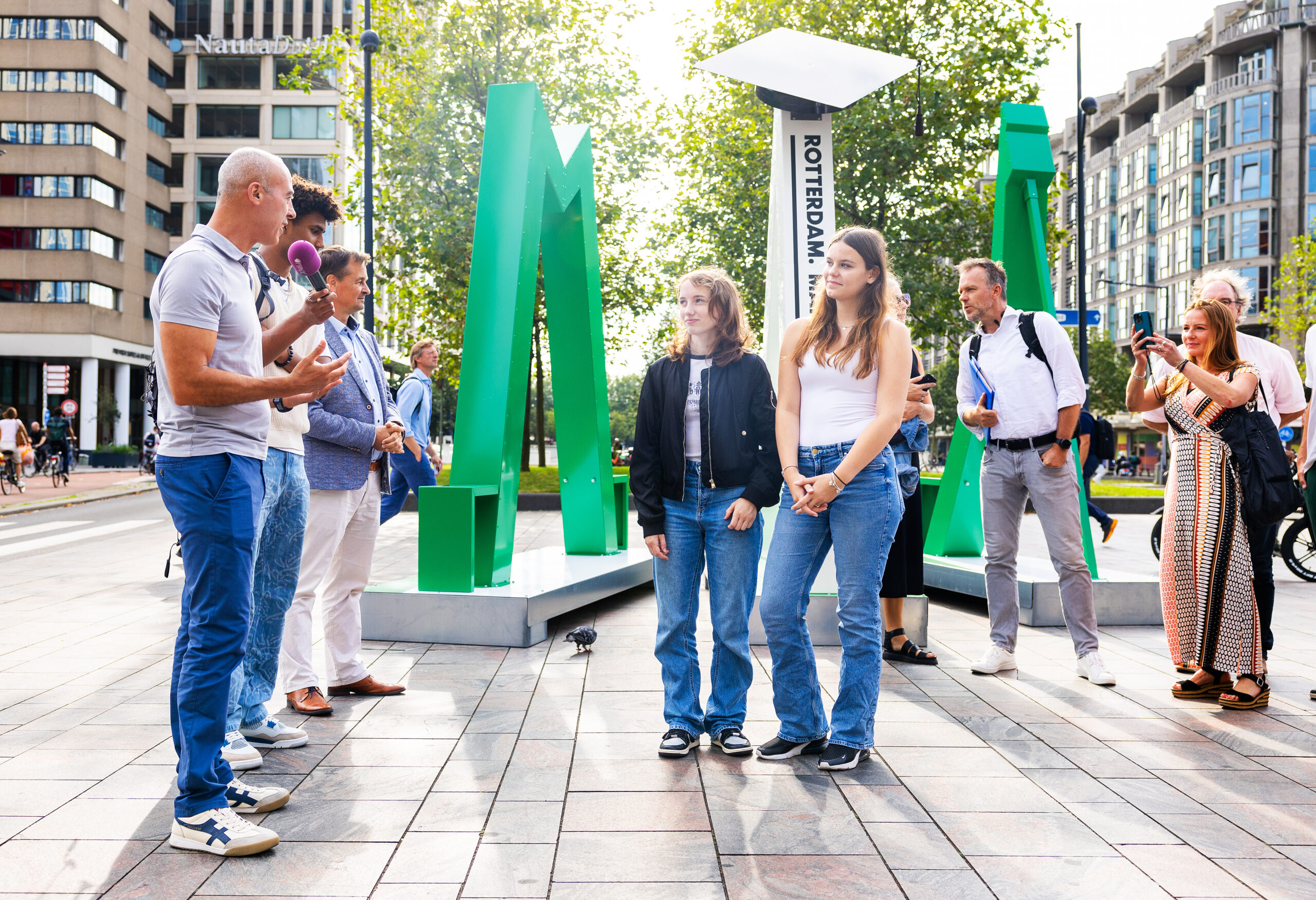 Mensen staan voor groene sculpturen in een stedelijke omgeving, met gebouwen op de achtergrond.