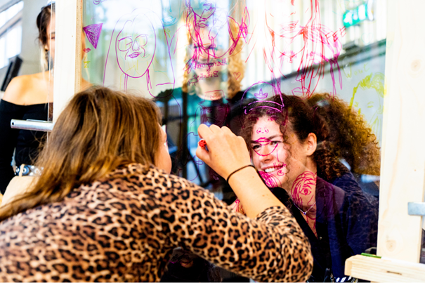Twee personen tekenen met stiften op een doorzichtige wand vol kleurrijke schetsen.