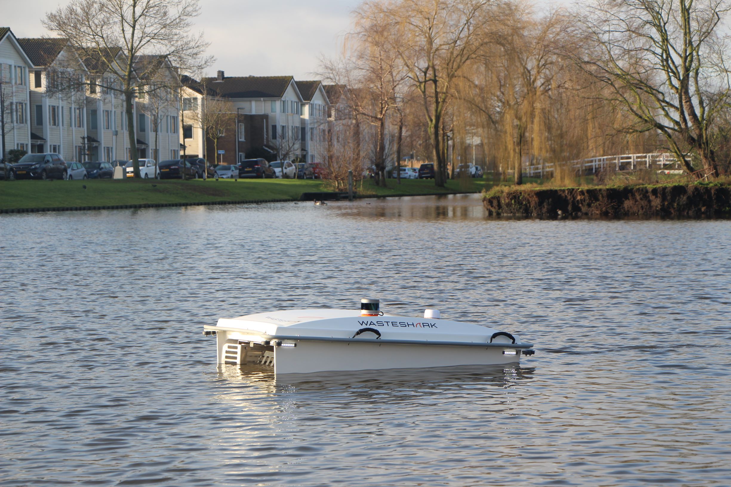 Een WasteShark-drone vaart op een waterweg met huizen en bomen op de achtergrond.