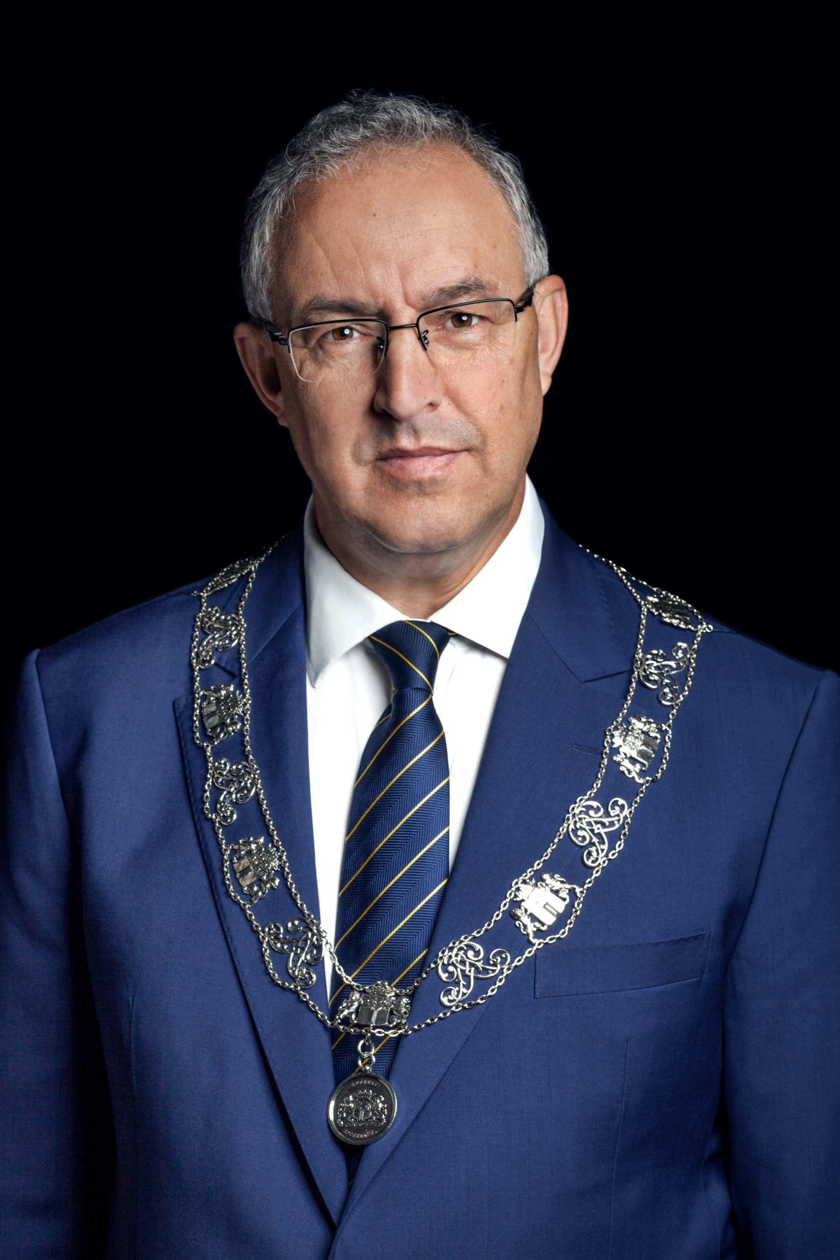Man in blauw pak met ambtsketen en bril tegen zwarte achtergrond