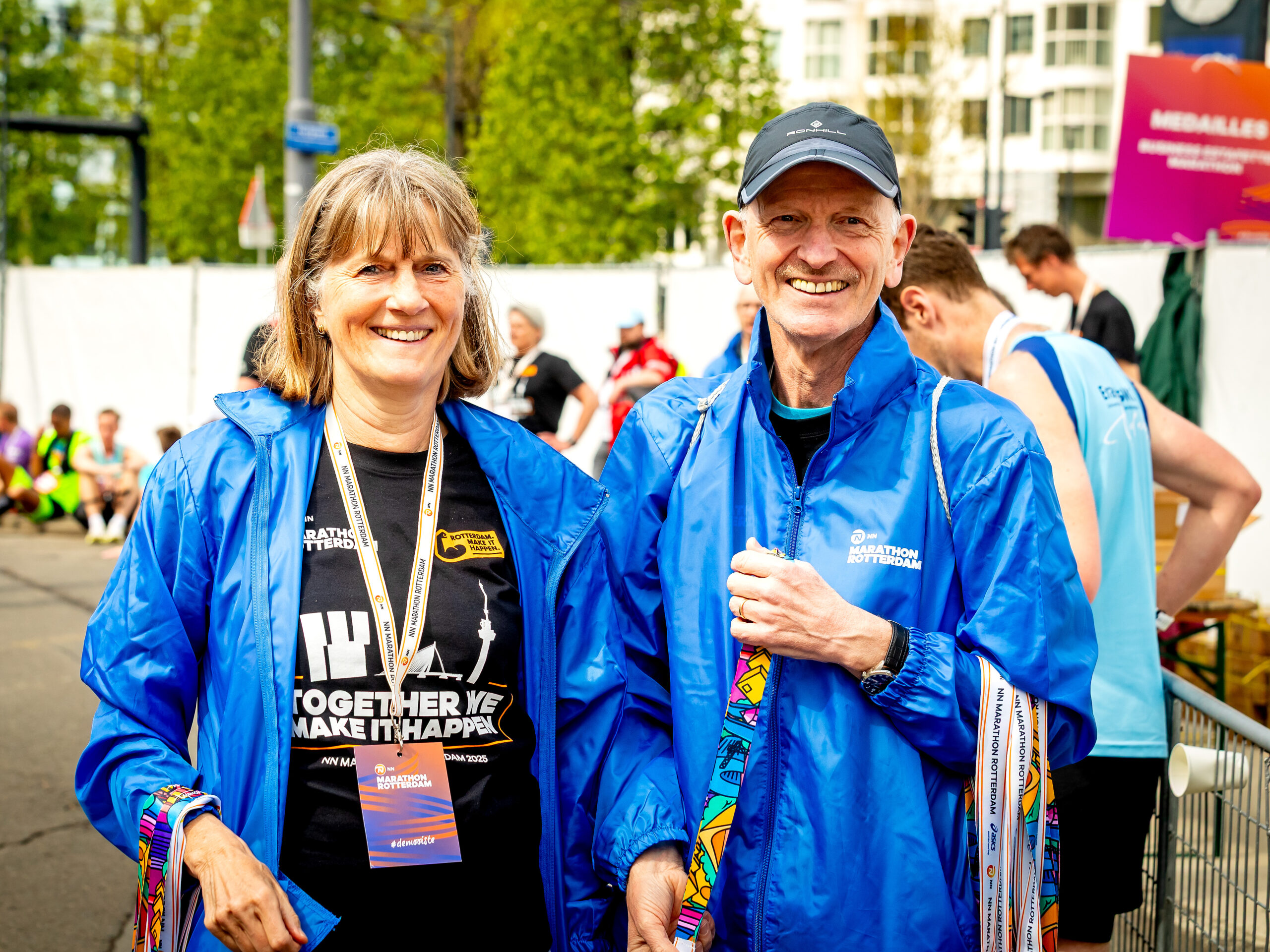Twee lachende mensen in blauwe jassen bij de Rotterdam Marathon, met medailles in hun handen.