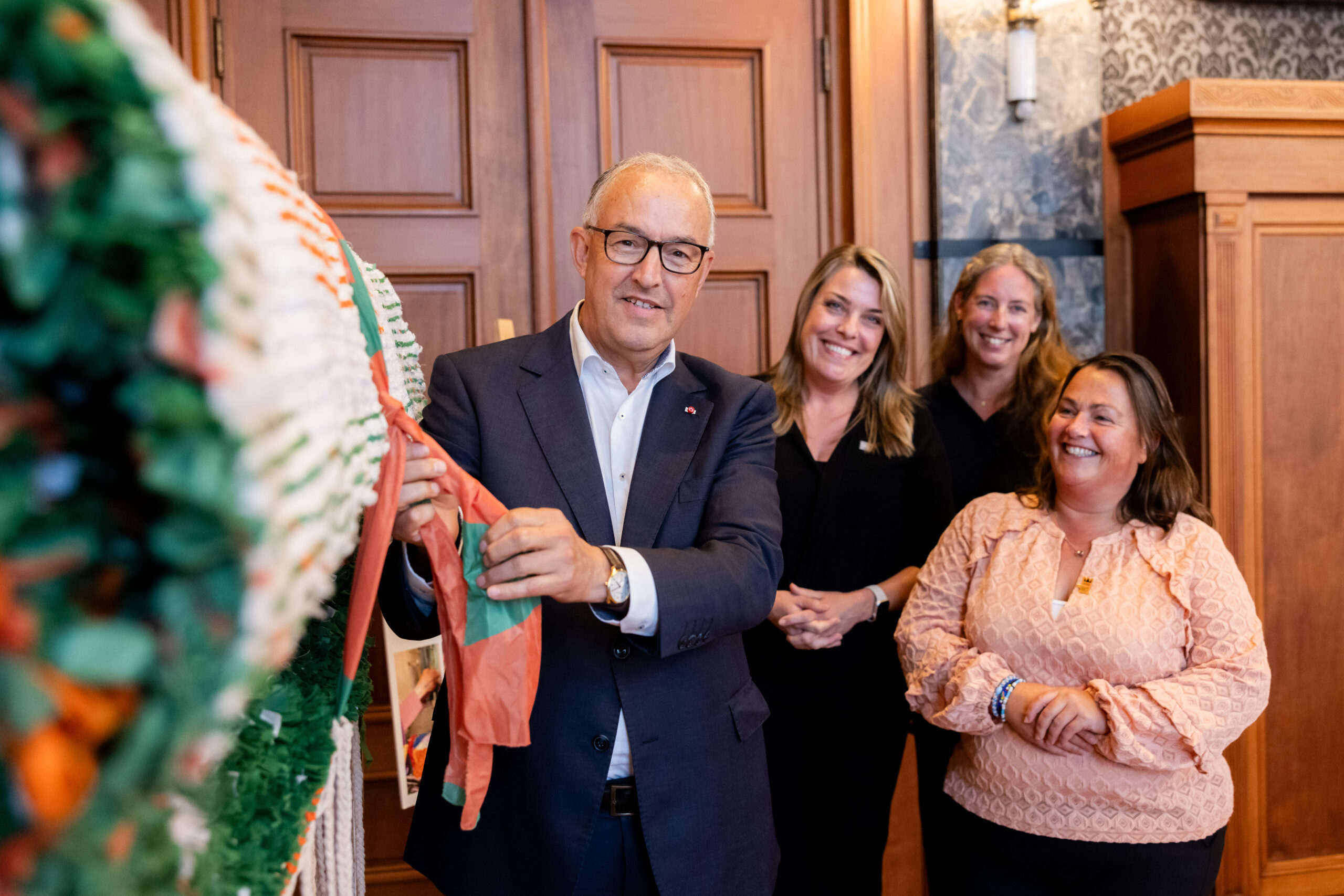 Een man in pak houdt een kleurrijk lint vast, met lachende vrouwen op de achtergrond.