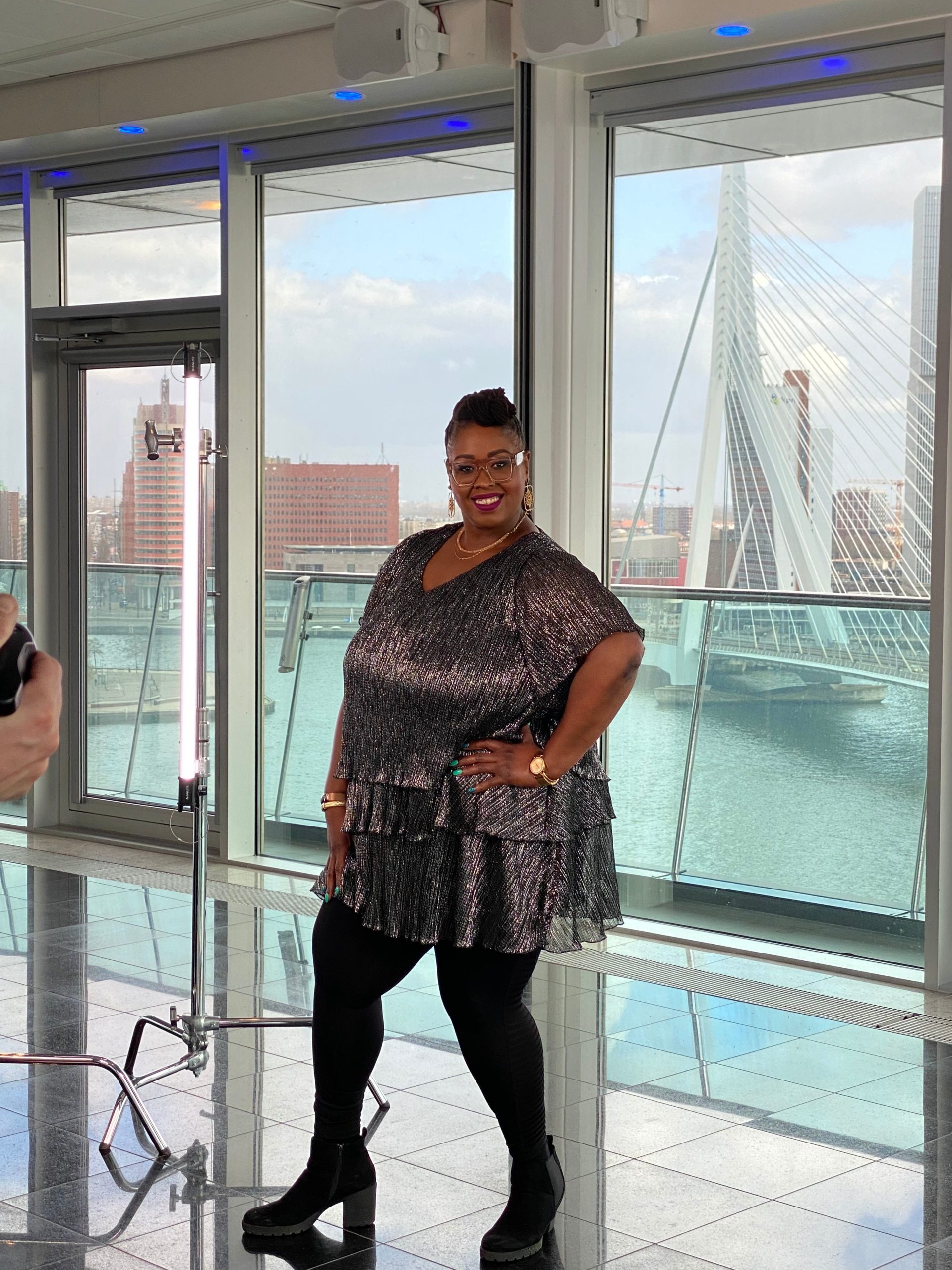 Vrouw poseert voor grote ramen met uitzicht op de Erasmusbrug in Rotterdam.
