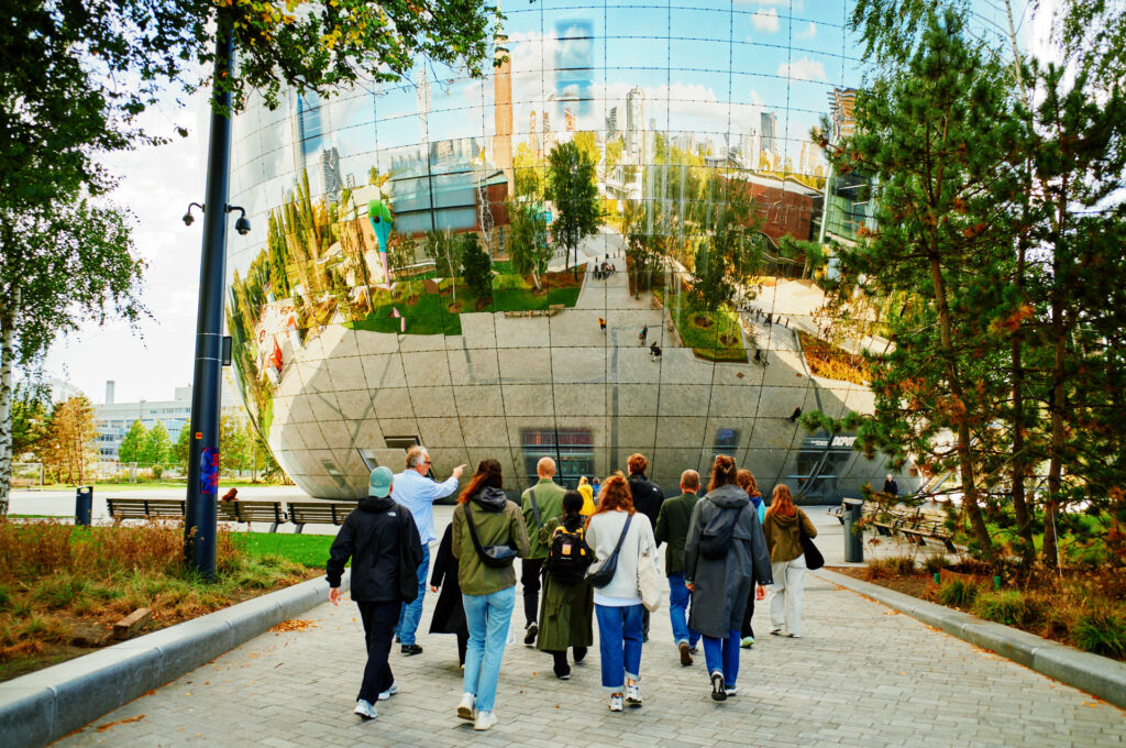 Een groep mensen loopt richting Het Depot van Museum Boijmans Van Beuningen. In de weerspiegeling van Het Depot zie je de stad op de achtergrond. 