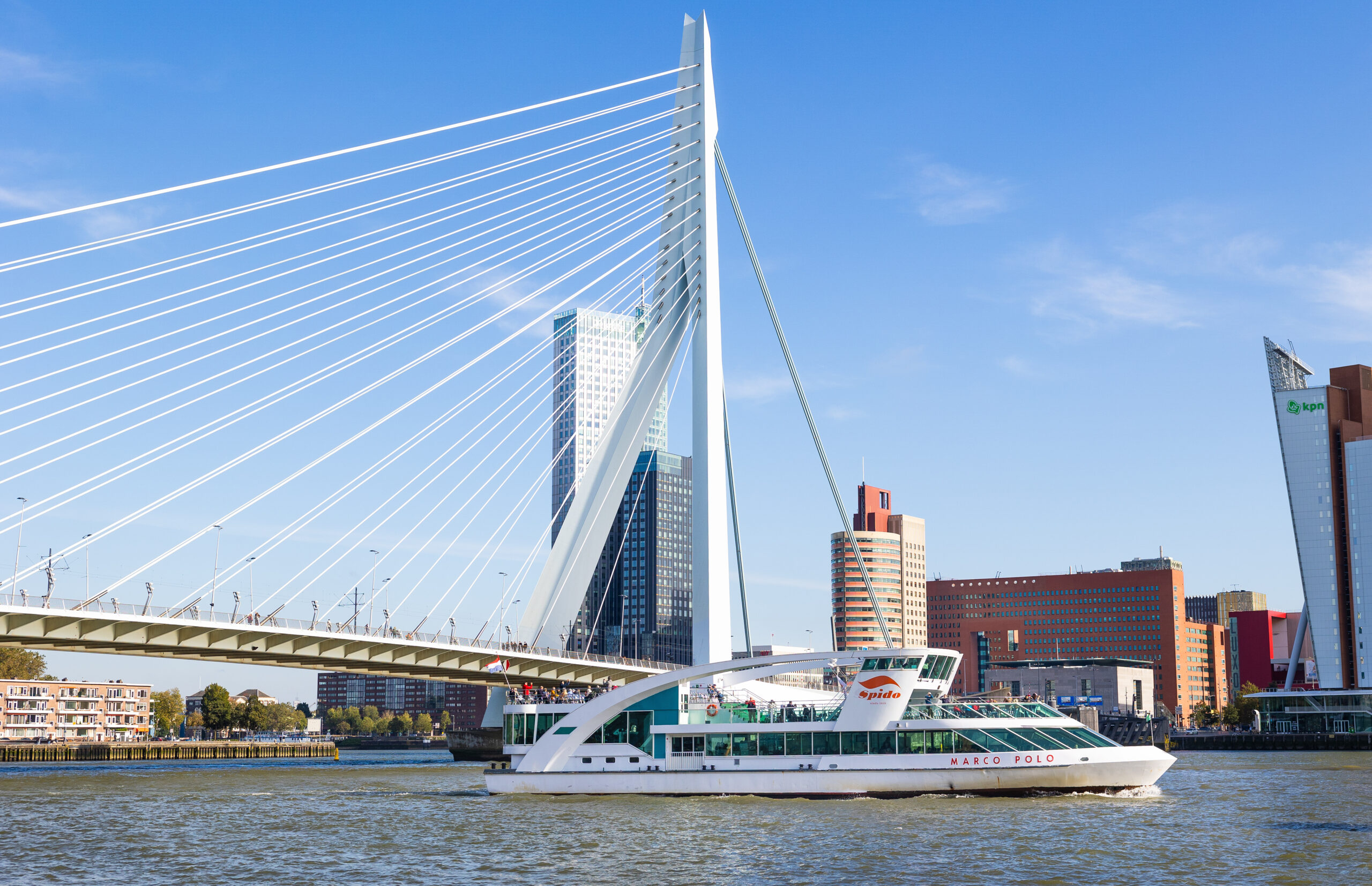 Een foto van de iconische Erasmusbrug en een Spido-boot die over de Maas vaart. Op de foto zie je ook de zuidkant van de stad met onder andere het Luxor theater, het kantoorgebouw van KPN en het kantoor van Deloitte.