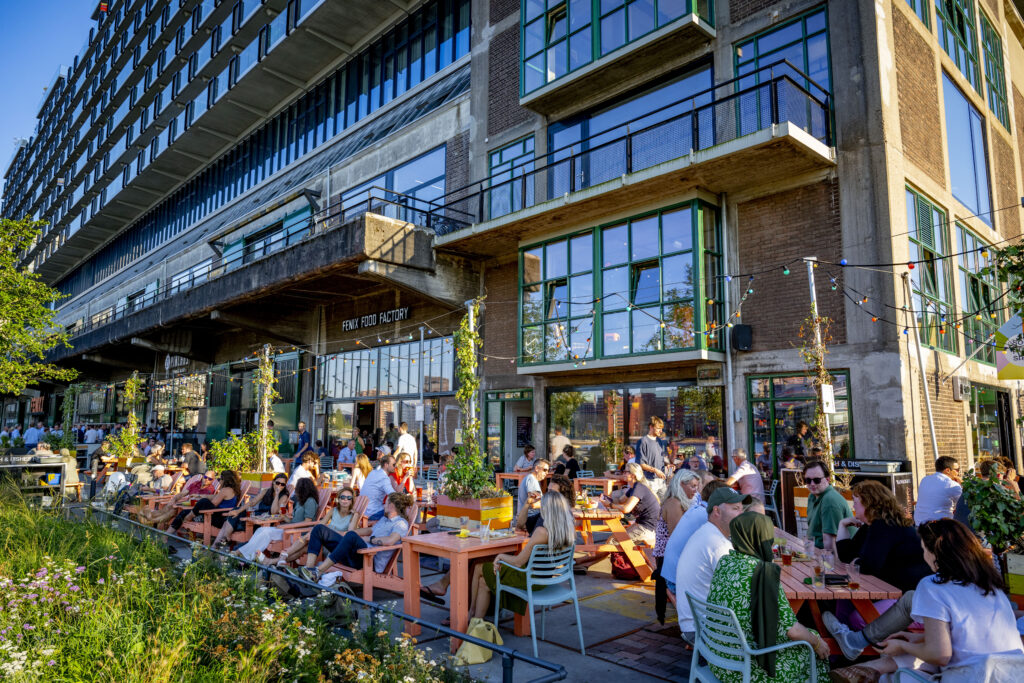Een foto van mensen die genieten van een zomeravond op de Kop van Zuid, bij de Fenix ​​Food Factory - een culinair centrum met een foodhall, bijzondere brouwerij en kraampjes voor vlees, groenten en zuivel.